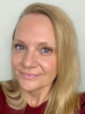 Woman with long blonde hair and blue eyes, smiling slightly, wearing a maroon top, in front of a plain background.