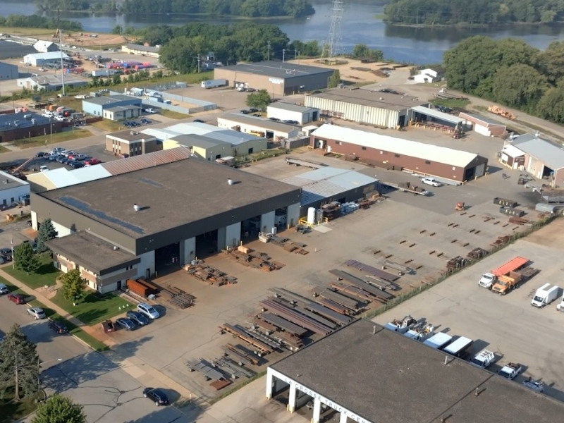 Aerial view of an industrial complex with warehouses, parked vehicles, and stacks of metal beams outside.