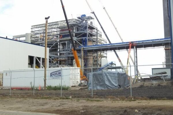 A construction site with cranes and scaffolding around a large industrial building, fenced off in the foreground.