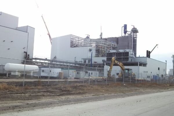 A large industrial facility with cranes and construction equipment near a fenced perimeter under an overcast sky.