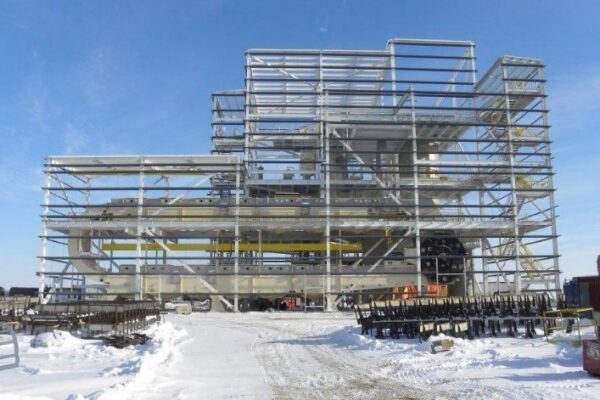 Steel framework of an industrial building under construction, surrounded by snow and equipment, under a blue sky.