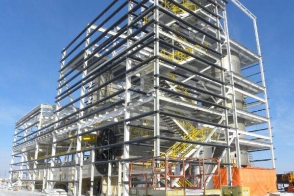 Steel industrial building frame under construction on a snowy site, with visible stairs and equipment nearby under clear blue sky.