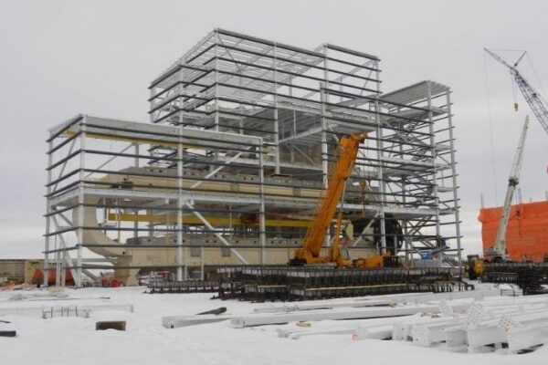 Steel framework of a large industrial building under construction in a snowy area, with cranes and equipment nearby.