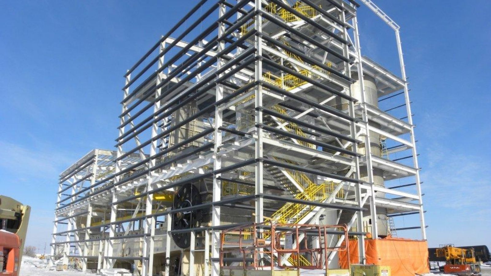 Steel-framed industrial building under construction with yellow stairs and equipment visible, set against a clear blue sky.