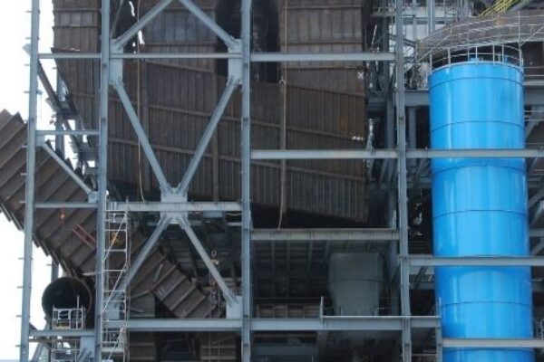 A blue Genie GTH-1056 lift in front of a large metal construction site with scaffolding and a tall blue cylinder.