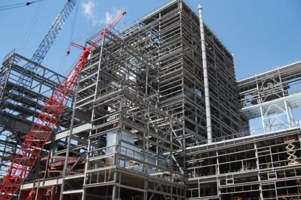 Steel framework of a large building under construction with cranes against a clear blue sky.