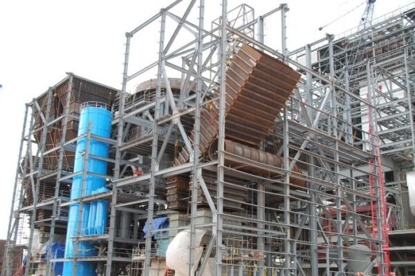 Large industrial structure under construction with metal framework, cranes, and various machinery visible on site.