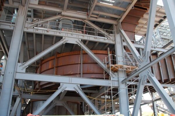 Steel framework and rust-colored curved metal structure under construction, with scaffolding and ladders visible.