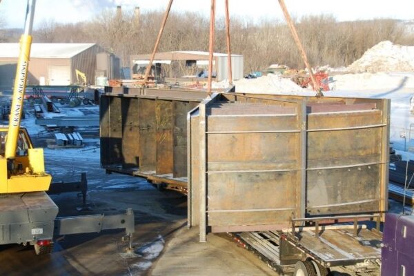 A large metal structure is lifted by a crane in an industrial outdoor area with snow on the ground.
