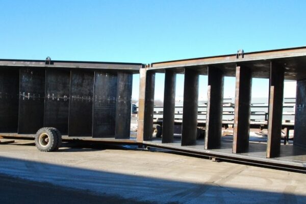 Large, empty steel box structure with open doors, sitting on a paved outdoor area under a clear blue sky.