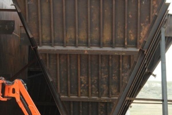 An orange boom lift is parked in front of a large, rusty, metal industrial hopper outdoors.