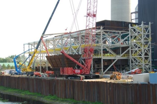 Construction site with cranes and steel framework by a body of water, heavy equipment and workers visible.