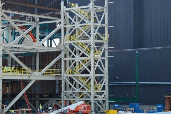 Steel building frame under construction with exposed beams, stairs, and workers in safety gear on multiple levels.