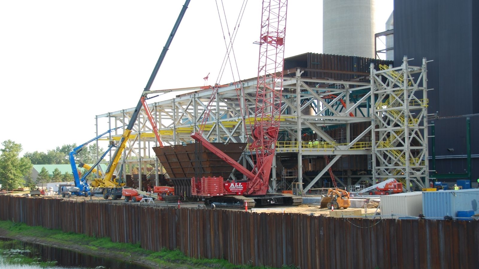 Construction site with cranes, steel framework, and heavy machinery beside a body of water.