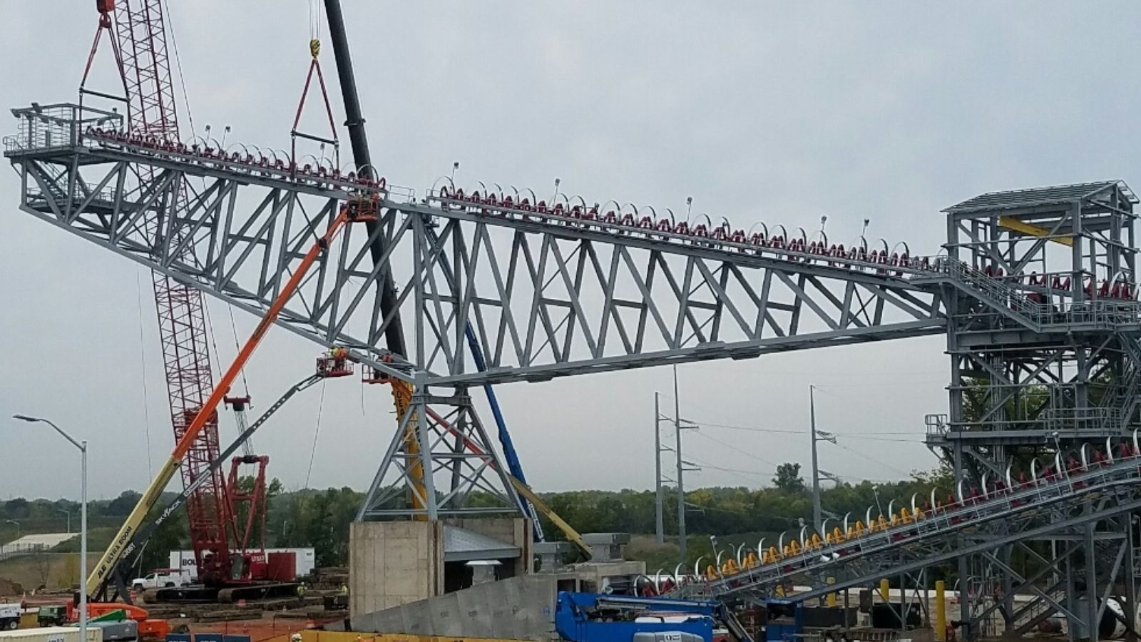 Large steel roller coaster track section being installed by cranes at a construction site under a cloudy sky.