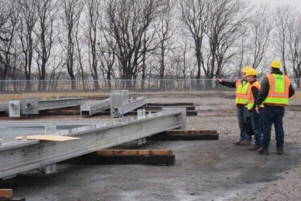 Three construction workers in safety gear stand near steel beams, with one pointing, at an outdoor worksite.