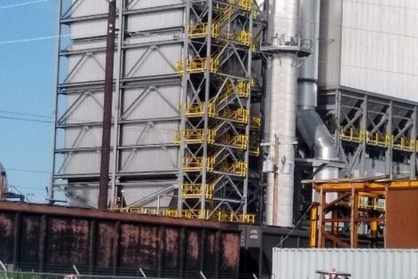 A large industrial building with metal pipes, stairs, and yellow railings, seen from a fenced area with construction materials.