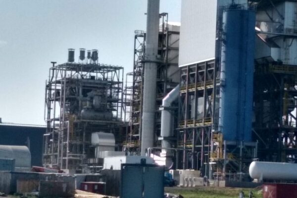 Industrial facility with large metal structures, pipes, and tanks, set against a blue sky with some equipment in the foreground.