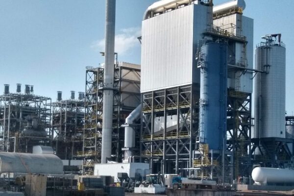 Industrial facility with large metal structures, storage tanks, and pipes under a clear blue sky.