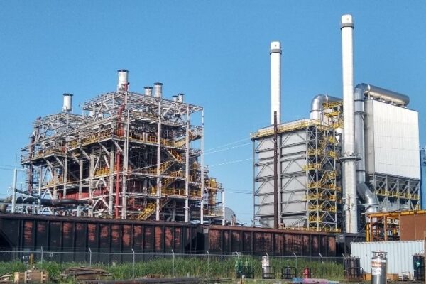 Industrial facility with large metal structures, pipes, and smokestacks under a clear blue sky.