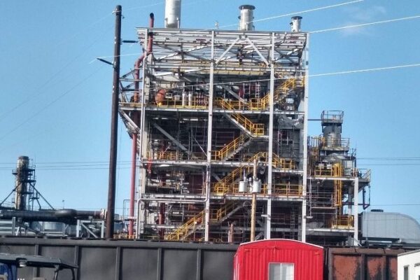 Industrial plant with metal framework, pipes, and stairs under a clear blue sky; red container in front.