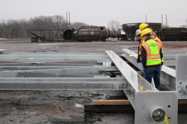 Three construction workers in safety gear inspect large steel beams at an industrial site with debris in the background.