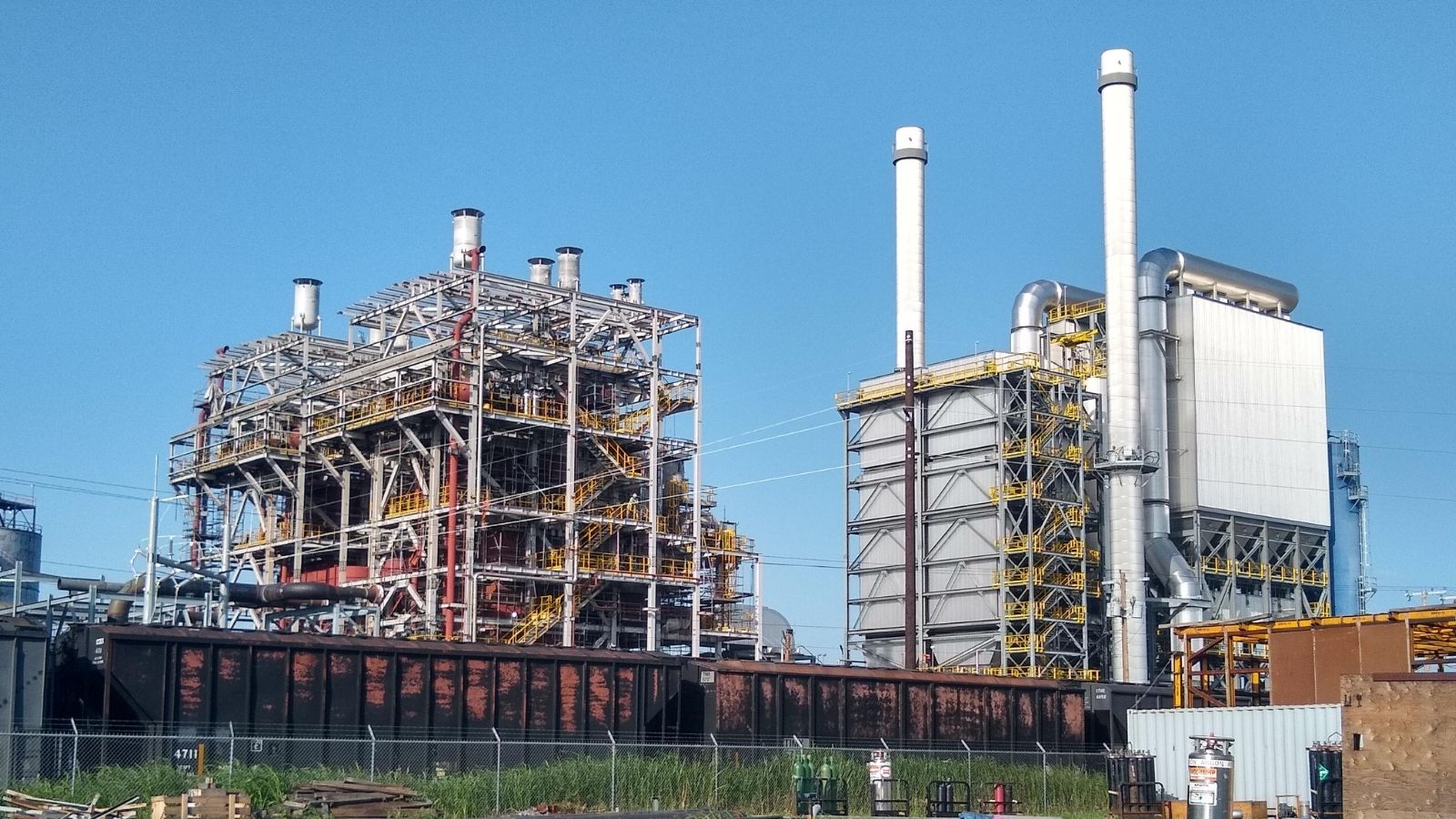 Two large industrial buildings with metal frameworks and tall smokestacks under a clear blue sky.