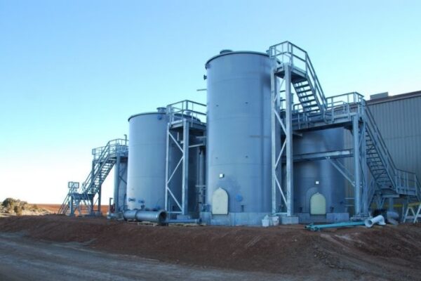 mid-city-steel-cow-canyon-central-processing-facility-industrial-gallery-3 Three large metal industrial tanks with staircases and platforms beside a gray building on a dirt ground under a clear sky.