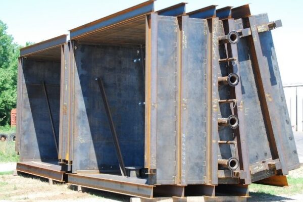 Large steel industrial structure with angled supports and pipe fittings, situated outdoors on a grassy area near a building.