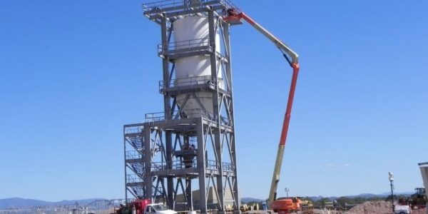 mid-city-steel-drake-cement-industrial-gallery-13 A worker on a cherry picker inspects a tall industrial metal tower at a construction site under a clear blue sky.