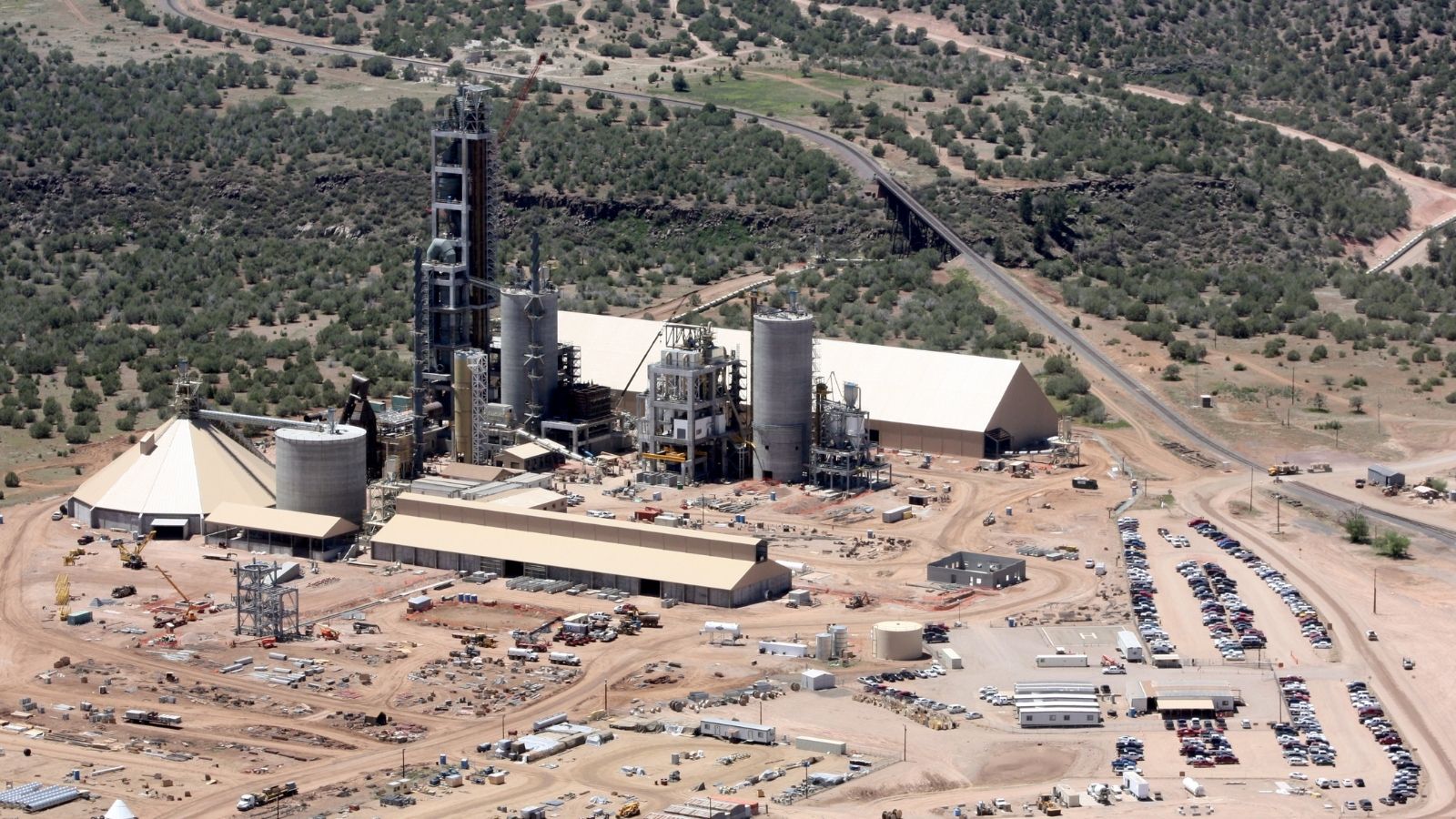 Aerial view of an industrial cement plant surrounded by parking lots, trees, and access roads in a rural area.