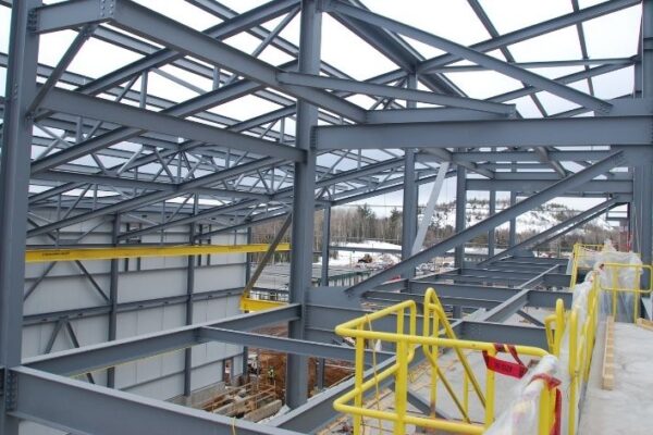 Steel framework of a building under construction with yellow safety railings and a snowy landscape in the background.