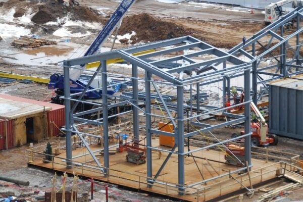 Steel building frame under construction, surrounded by equipment, materials, and workers at a muddy worksite.
