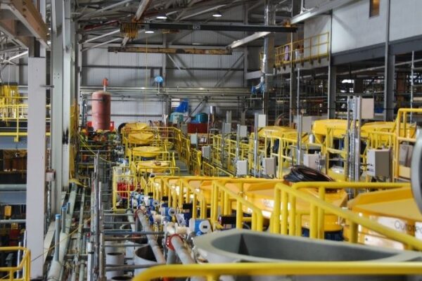 Factory interior with rows of yellow machinery, metal pipes, and industrial equipment in a spacious, well-lit environment.