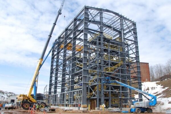 Large steel-frame building under construction with cranes, surrounded by snow and some workers on the ground.