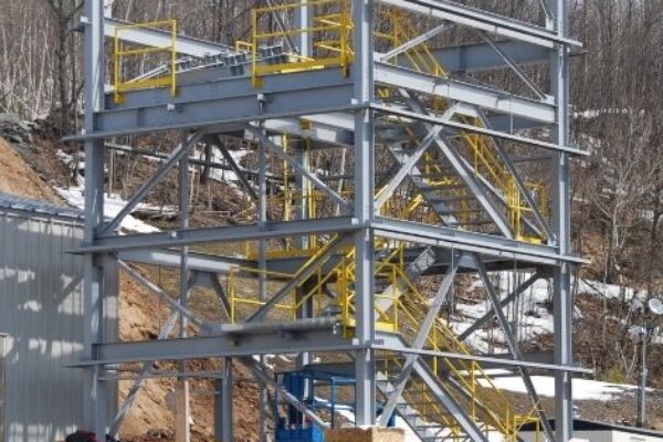 A steel framework of a building under construction, with yellow railings and a dirt ground, set against trees.