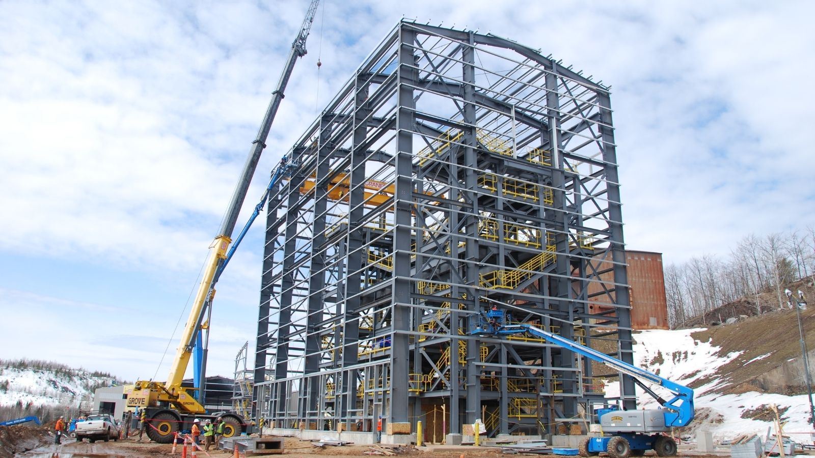 Large steel framework of a building under construction with cranes and workers on a snowy site.