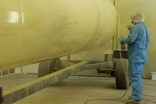 Person in blue coveralls spray-painting a large cylindrical tank yellow in an indoor facility.