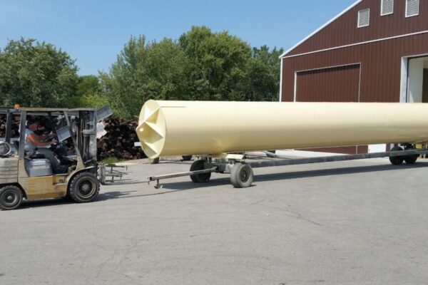 A forklift moves a large, light yellow cylindrical tank on a wheeled trailer outside a brown industrial building.