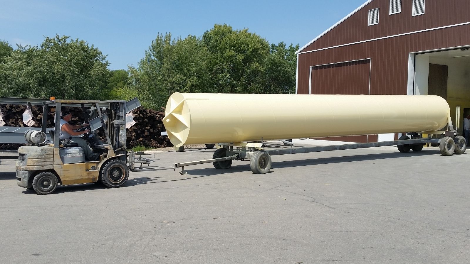 A forklift moves a large yellow cylindrical tank on a wheeled cart outside a brown industrial building.