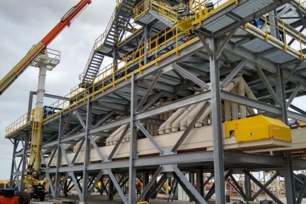 Large industrial structure with yellow railings, stairs, and workers nearby; construction equipment is present.