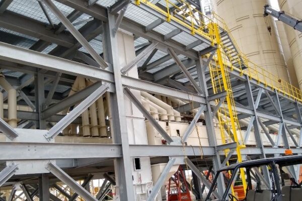 Steel industrial structure with pipes, yellow safety railings, and a forklift in a factory or plant setting.