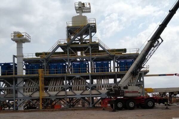A construction crane works at an industrial site with metal structures and equipment under a cloudy sky.