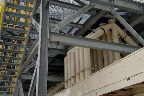 Industrial structure with metal stairs and pipes; worker in safety gear stands below on a concrete floor.