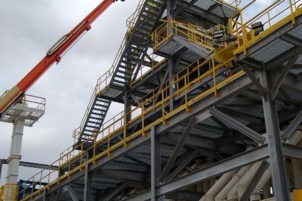A crane lifts a worker in a bucket to the top of an industrial structure with stairs and yellow railings.