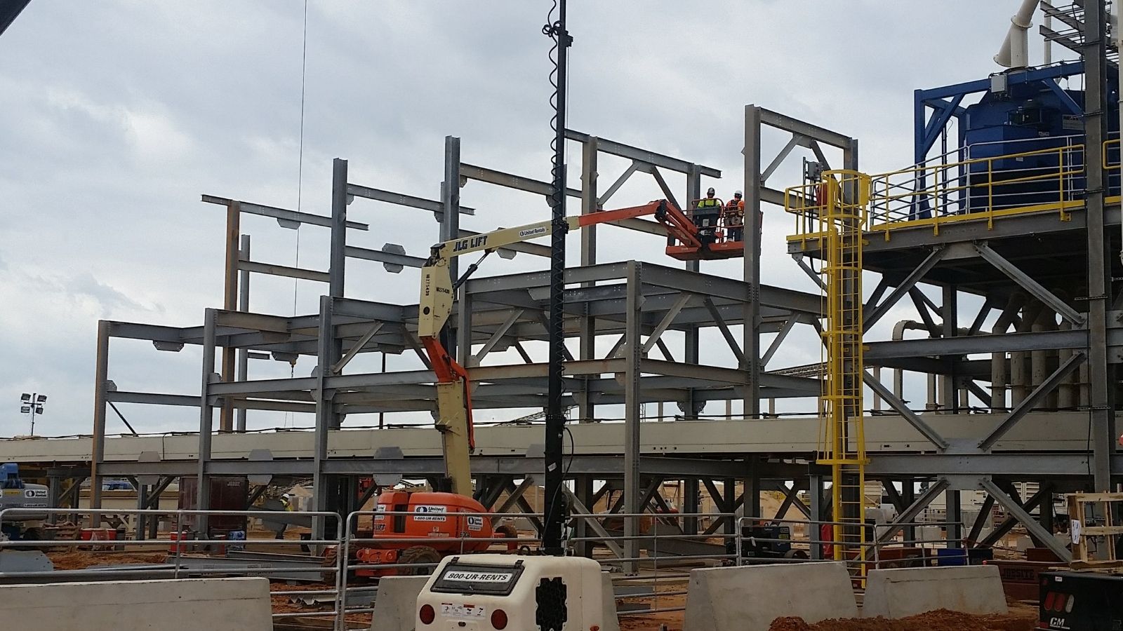 Construction workers using a lift to assemble steel framework on a large building site with machinery nearby.