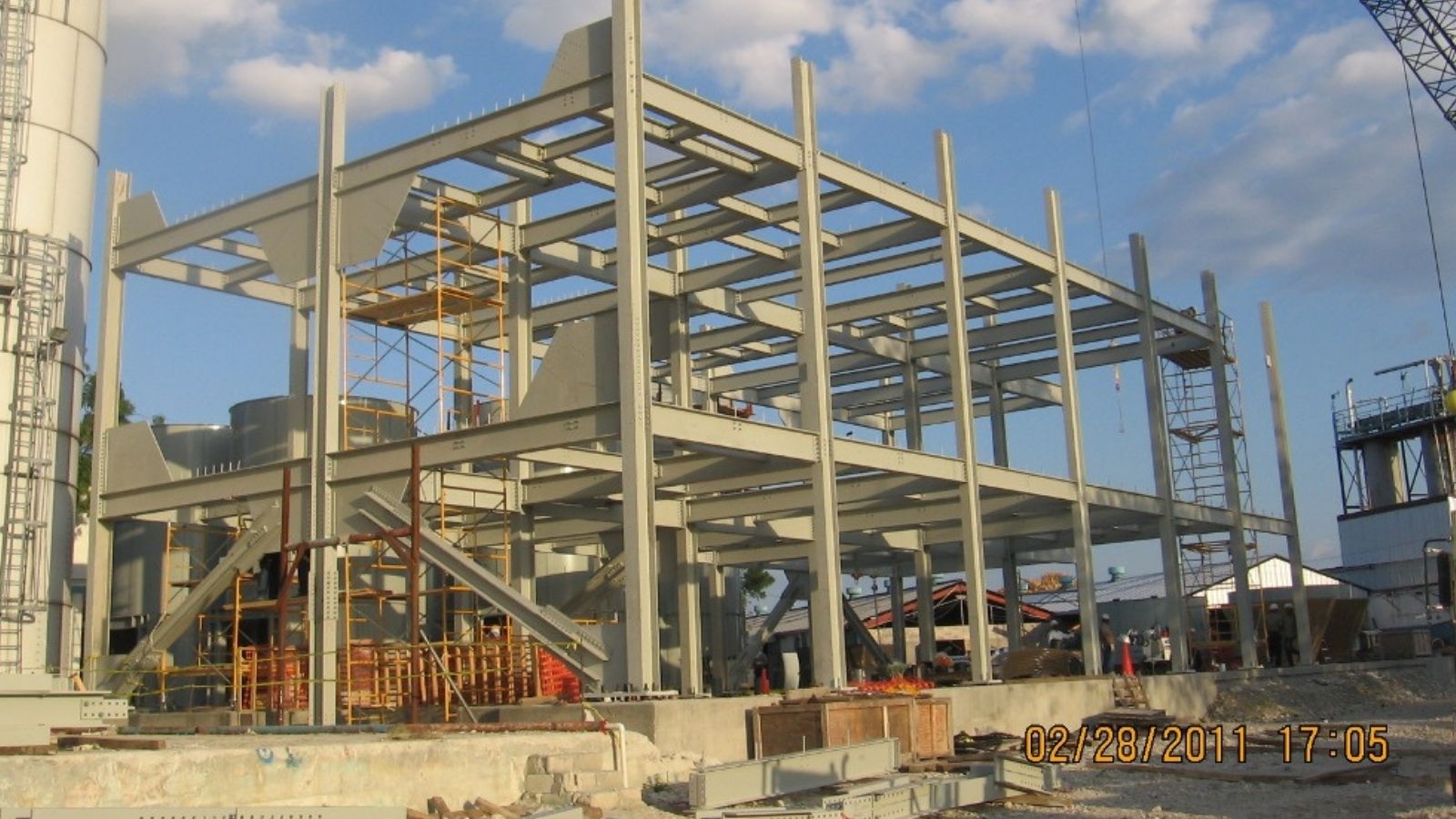 Steel frame structure of a building under construction, with scaffolding and equipment on site, against a blue sky.