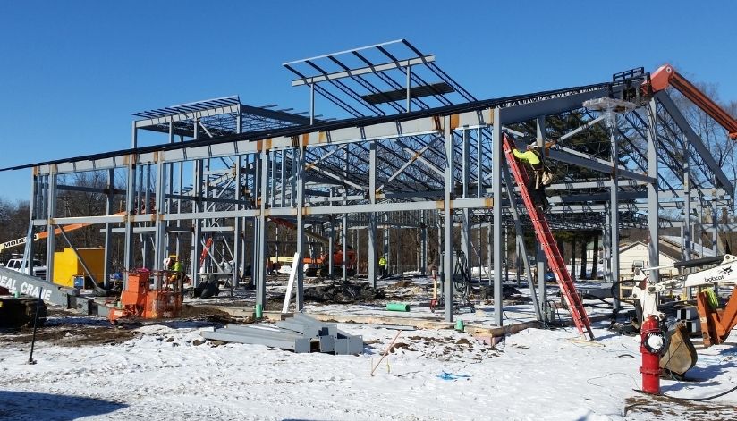 mid-city-steel-holmen-library-commercial-project-gallery-1 Steel framework of a building under construction on a snowy ground, with workers and construction equipment visible.