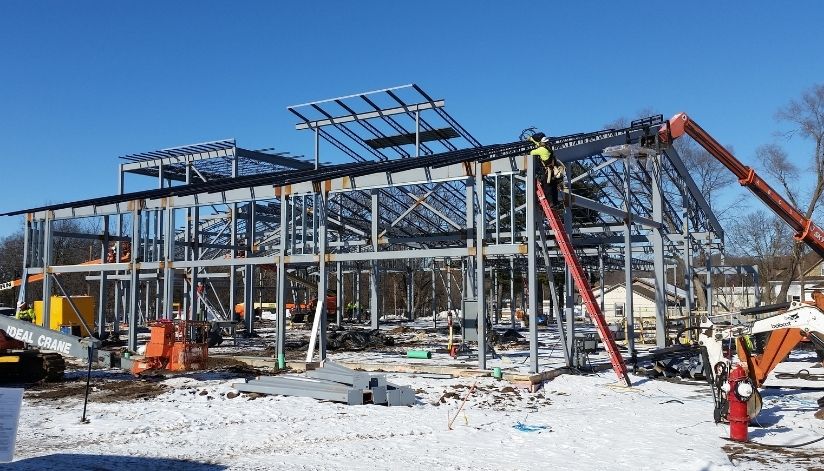 mid-city-steel-holmen-library-commercial-project-gallery-2 Steel framework of a building under construction, with workers and equipment on a snowy ground under a clear blue sky.