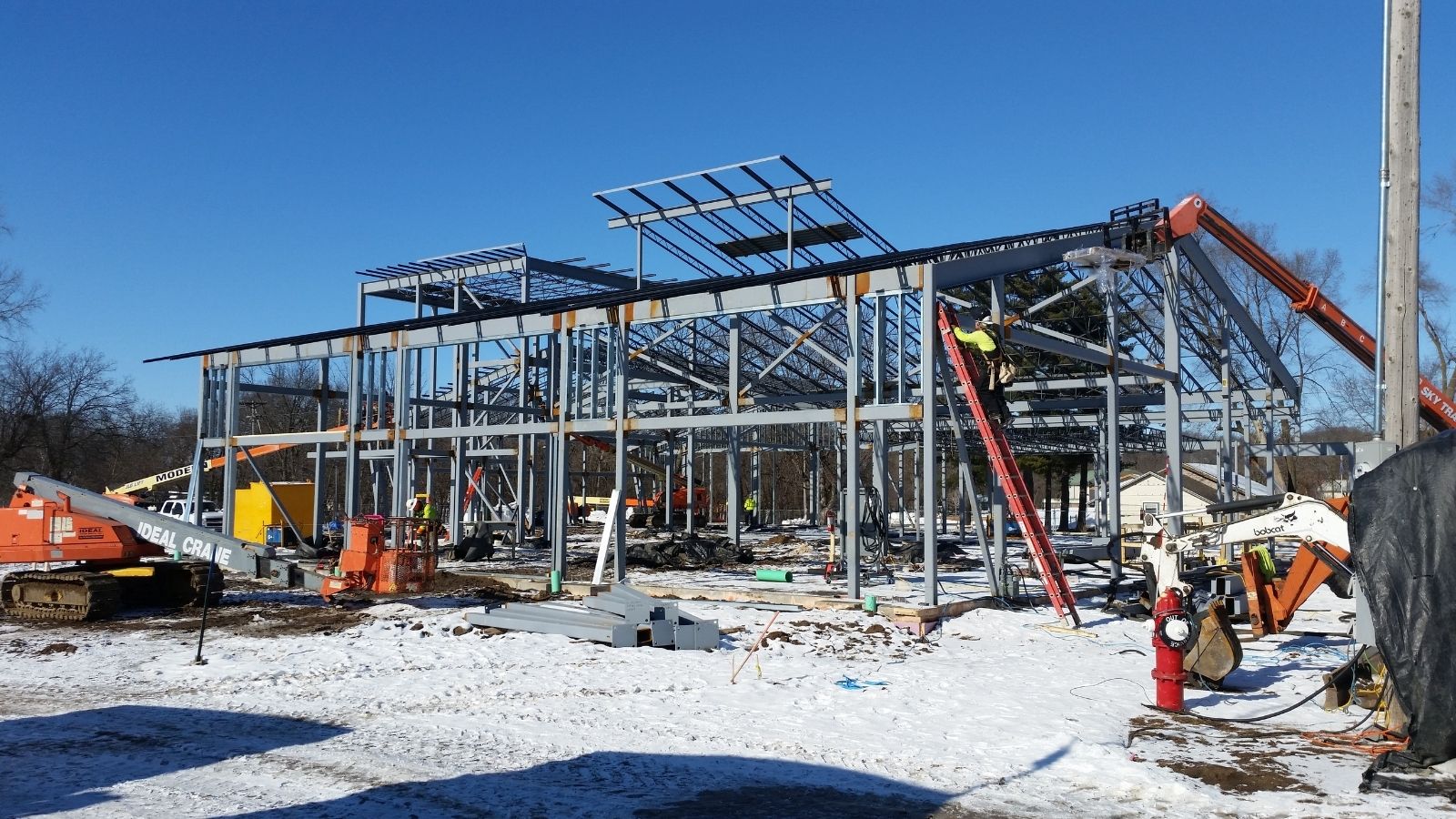 A steel building frame under construction with workers and equipment on a snowy ground under a clear blue sky.